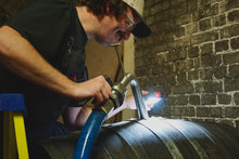 Load image into Gallery viewer, Person working on a barrel with tools in a dimly lit room.
