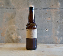 Load image into Gallery viewer, Beer bottle labeled 'The Kernel' on a wooden surface with a concrete wall background
