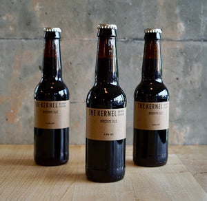Multiple 330ml dark glass bottles of Brown Ale with a light brown label sat upon a wooden table infront of a rustic cement wall. 