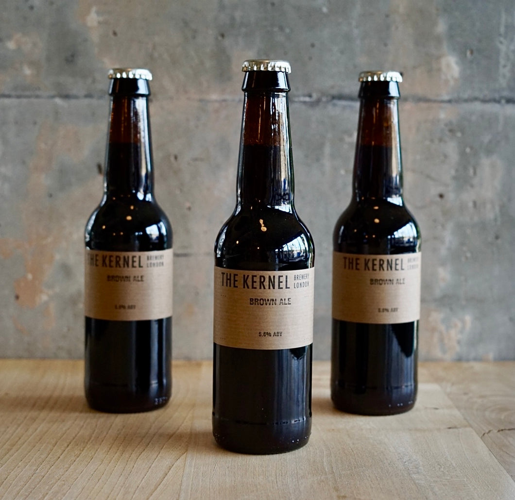 Multiple 330ml dark glass bottles of Brown Ale with a light brown label sat upon a wooden table infront of a rustic cement wall. 