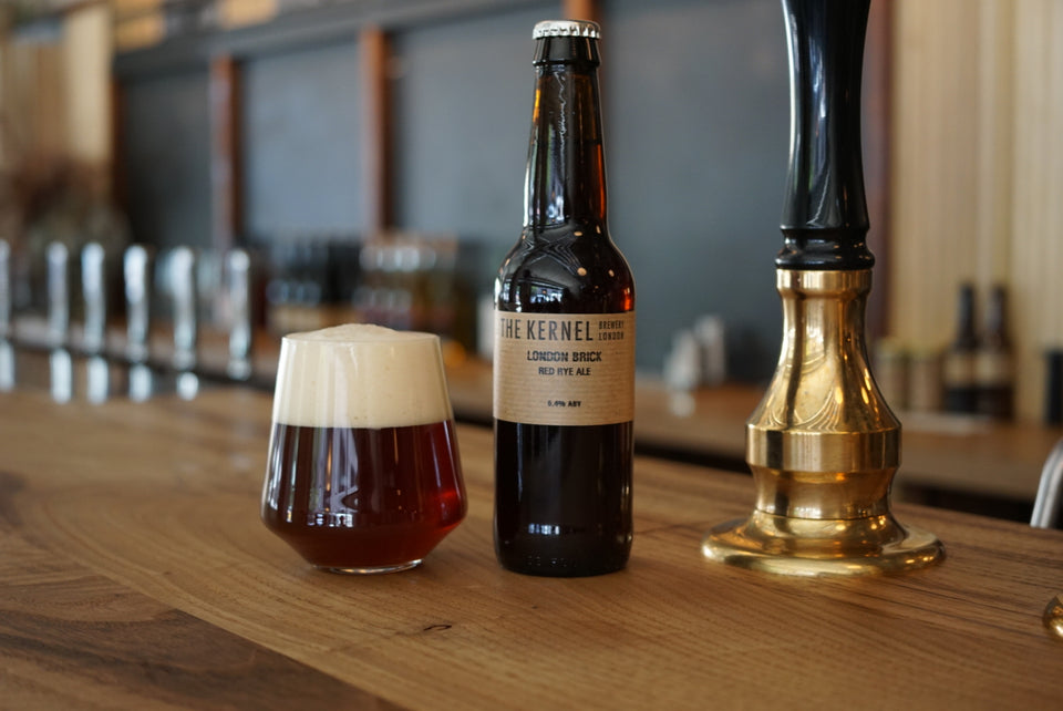 A single 330ml dark glass bottle of London Brick, with a light brown label is stood to the right of a schooner full of dark ruby beer with a very froathy head. Both are sat upon a bar top next to cask pump handle. 