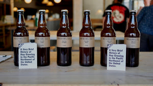 Six 500ml dark glass bottles of Pale Ale with various hops, each has a light brown label and is sitting upon a wooden table in a taproom. There are two small booklets infront of the bottles. 