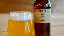 Load image into Gallery viewer, Bottle of 'The Kernel' Pils Saphir beer next to a glass of beer on a wooden surface.
