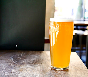 A pint glass full of East India pale Ale which is a vibrant golden heritage beer wth a foamy head, sat on a rustic wooden table with a dark walled background
