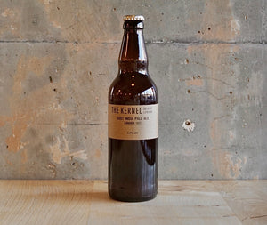 A 500ml dark glass bottle of East India Pale Ale with a light brown label set on a wooden table infront of a rustic cement wall. 