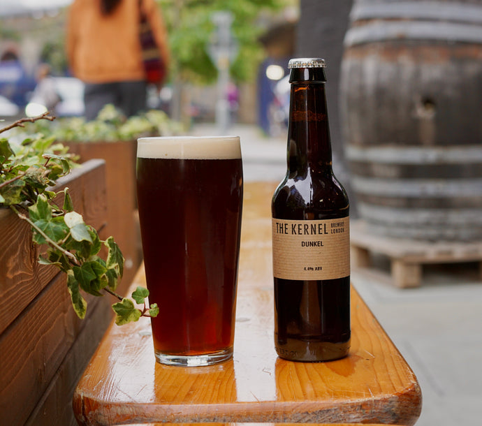 Bottle and glass of dark beer labeled 'The Kernel' on a wooden surface outdoors.